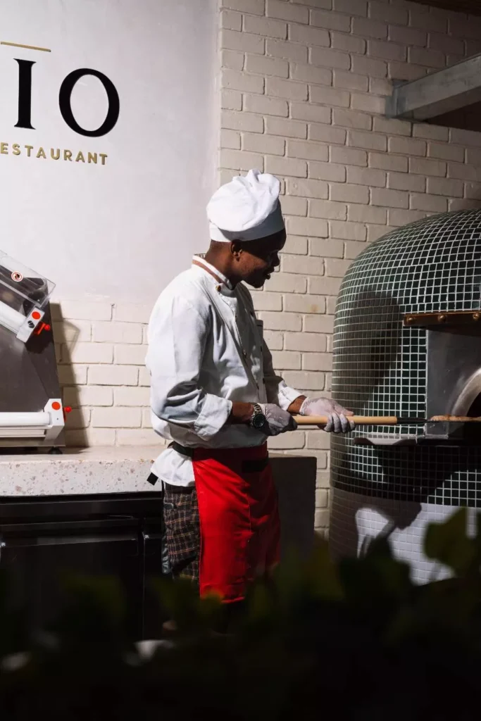Side view of a chef sliding pizza into a wood-fired oven with a long peel in a restaurant kitchen.