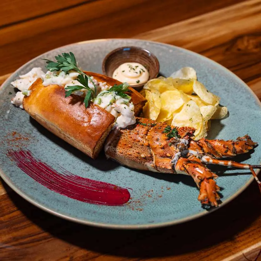 Close-up of a lobster roll filled with seafood, plated beside chips and a ramekin of dipping sauce.