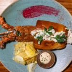 Lobster roll in a brioche bun served with potato chips, dip, and whole cooked lobster on a plate.