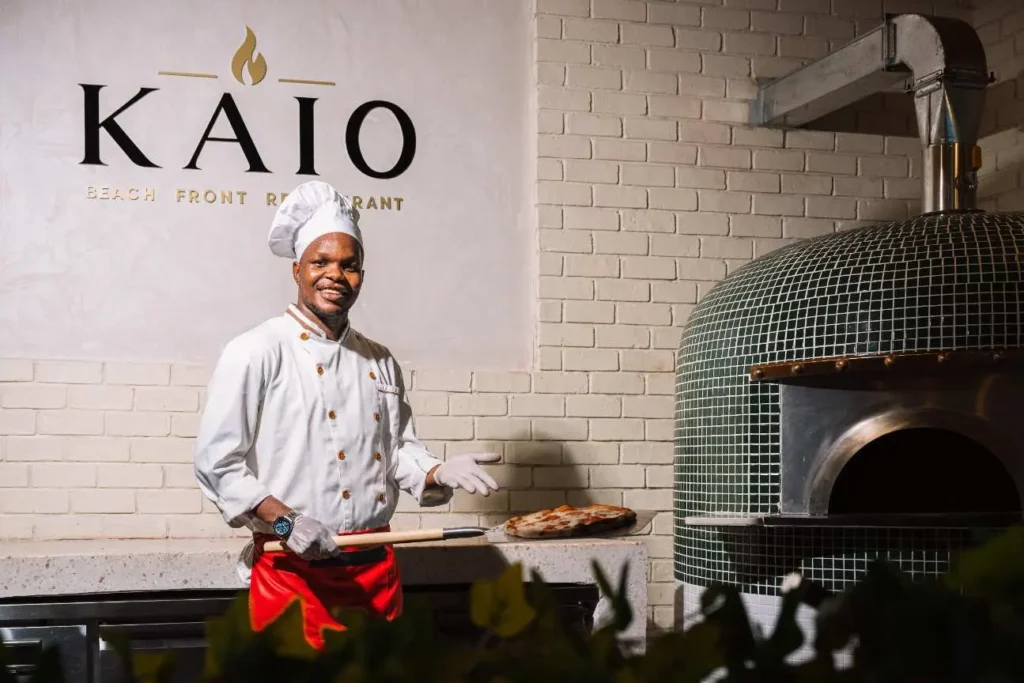 Professional pizza chef holding a fresh pizza next to a tiled oven at KAIO Beach Front.