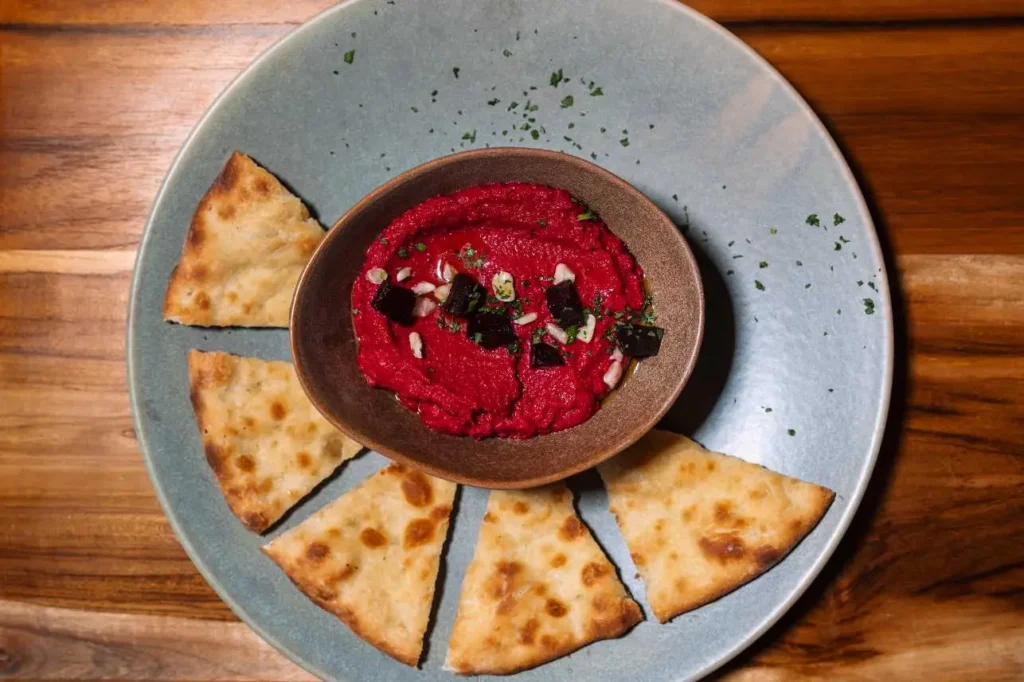 Smooth pink beetroot hummus garnished with herbs and served with baked crostini bread triangles.