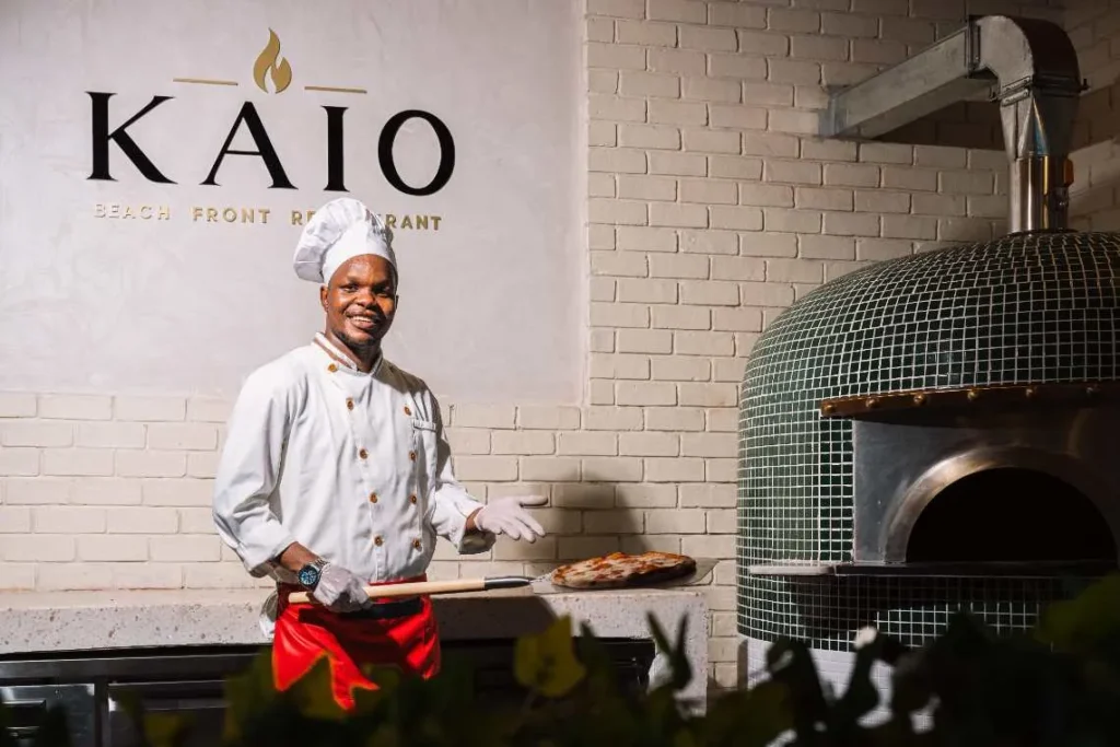 Chef holding a fresh pizza on a peel beside a green tiled wood-fired oven at KAIO Restaurant.