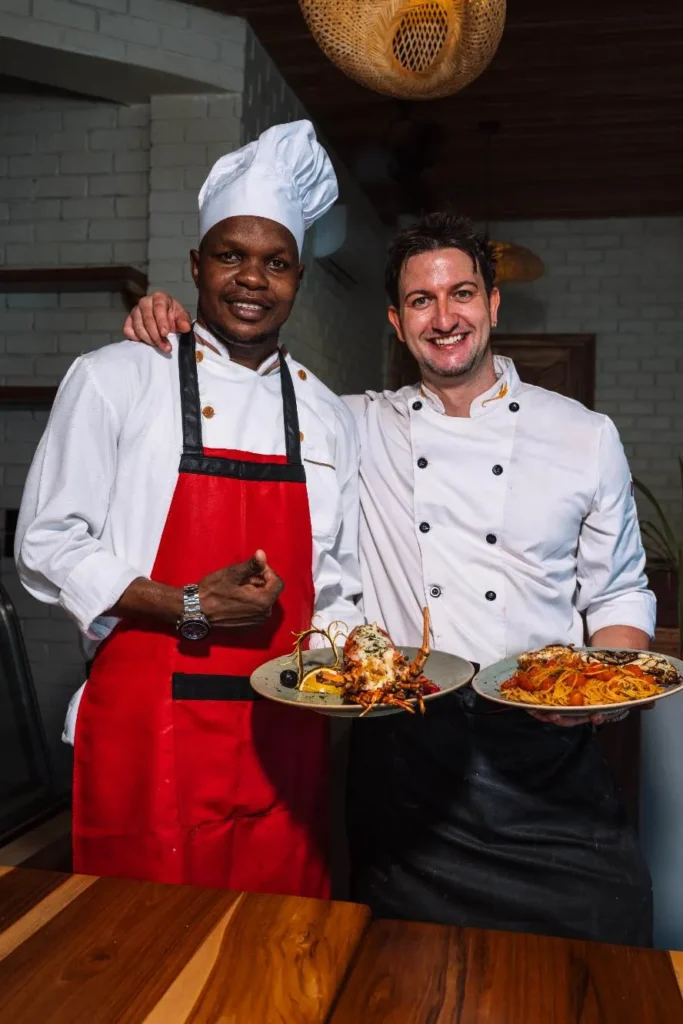 Two smiling restaurant chefs holding plates of grilled lobster and seafood pasta in a dining room.