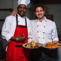 Two smiling restaurant chefs holding plates of grilled lobster and seafood pasta in a dining room.